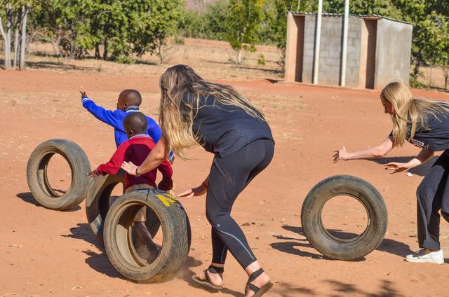 Two volunteers rolling tyres with a small child on red earth