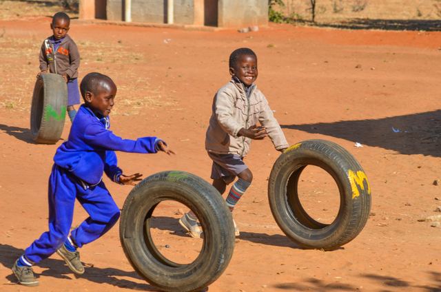 A boy in blue overalls running with a tyre while another child watches