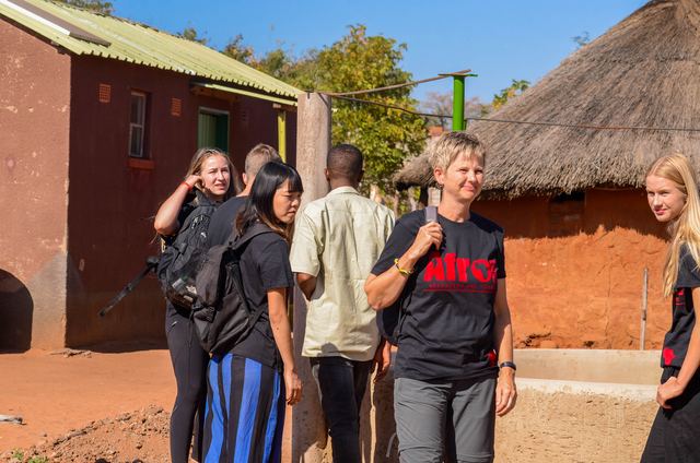 Volunteers and a local guide beside a mud-brick house with a thatched roof