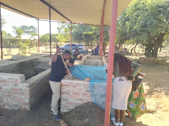 Two people working at a brick compost bay under a roofed shelter, with blue netting over the compost