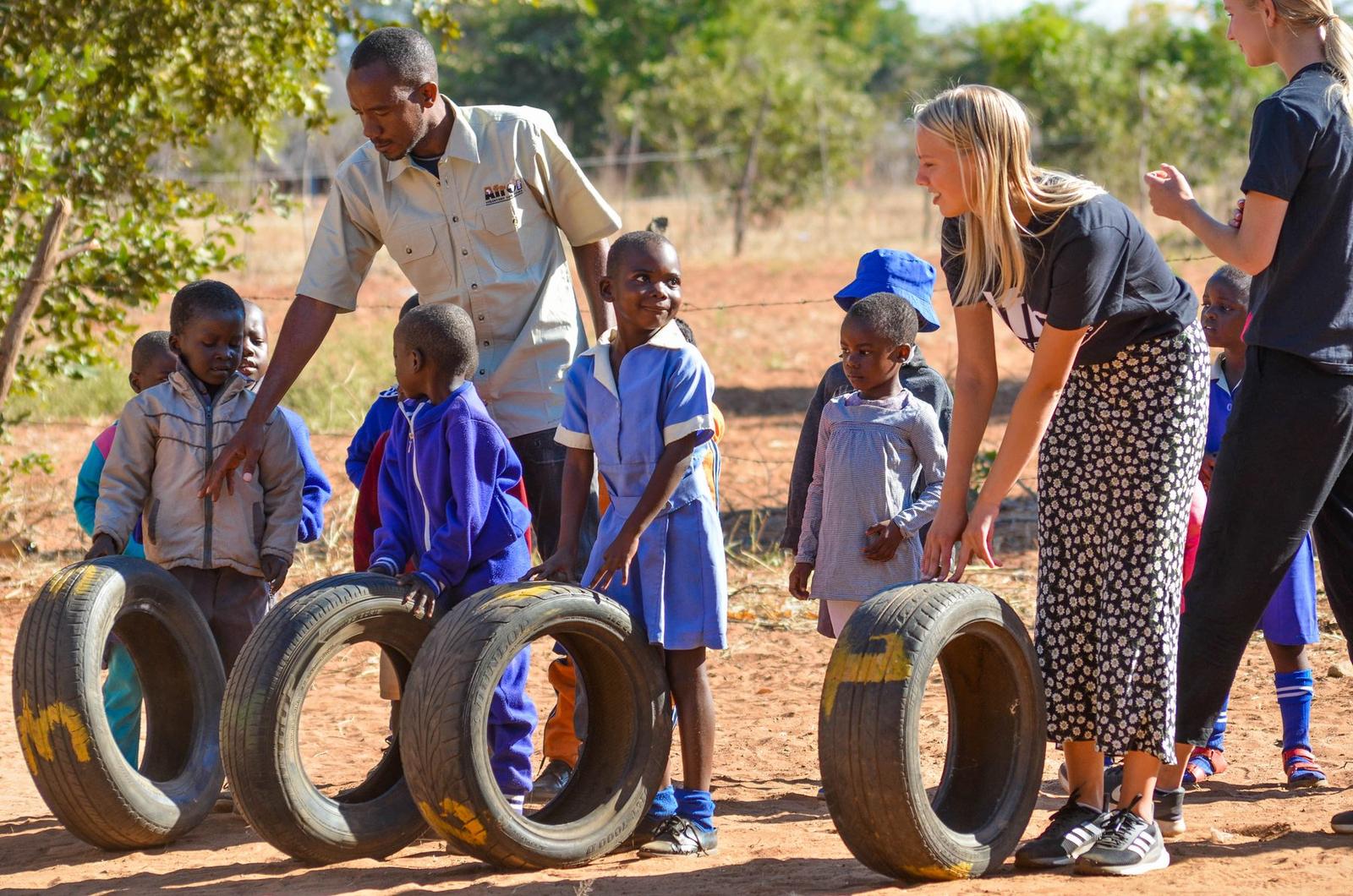 A volunteer and a community staff member playing a tyre-rolling game with children in school uniform on red earth